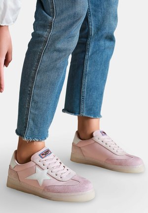 Personne portant des baskets rose clair avec un détail étoile blanche et un jean bleu aux ourlets effilochés sur fond blanc.