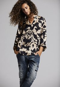 Schwarze und beige gemusterte Seidenbluse mit V-Ausschnitt, langen Ärmeln und lockerer Passform, kombiniert mit abgenutzten blauen Jeans.