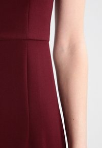 Bordeaux jurkstof met een gladde textuur, een getailleerd ontwerp en zichtbare naden. Inclusief een close-up van een arm tegen de jurk.