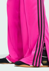 Roze sportbroek met zwarte strepen aan de zijkanten, met ritsen aan de manchetten. Zwart-witte schoenen met een tan kleurige zool zijn zichtbaar.