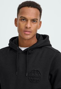 Juodas medvilnės džemperis su virvute, su toniniu apvaliu "NAVIGATE" logotipu ant krūtinės. Lygus paviršius, kasdieniška siluetas.