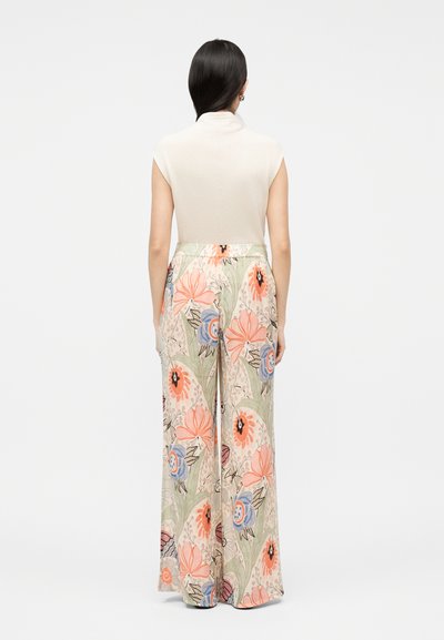 Γυναίκα με σκούρα μαλλιά, φοράει λευκό αμάνικο τοπ και φαρδιά παντελόνια με λουλουδάτο σχέδιο, στέκεται με φόντο απλό φωτεινό τοίχο, σε όψη από πίσω.