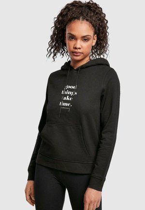 Schwarzes Hoodie aus weichem Material mit Kängurutasche, Kordeln und weißem Text "Gute Dinge brauchen Zeit" auf der Vorderseite.