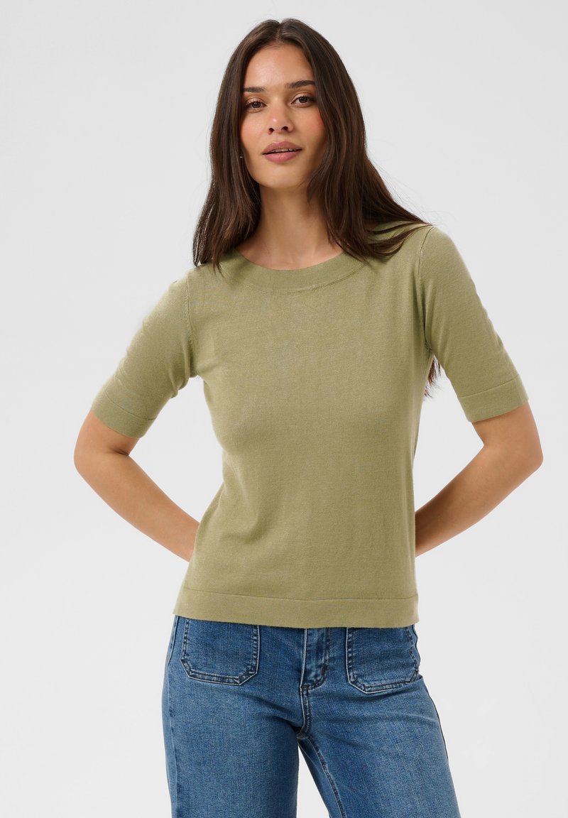 Femme aux longs cheveux bruns portant un pull léger vert clair à manches courtes et un jean bleu, debout les mains derrière le dos.