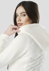 Fehér kapucnis pulóver nagy, puha kapucnival és steppelt, párnázott oldalsó panellel. Sima textúra enyhe fényű felülettel. A modell keze a galléron pihen.