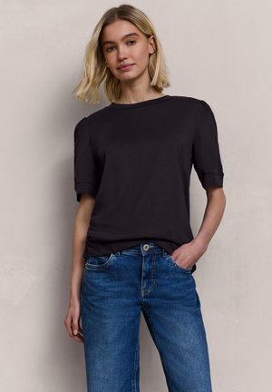 Junge Frau mit blondem Haar, die ein schwarzes kurzärmeliges Oberteil und blaue Jeans trägt, steht mit einer Hand in der Tasche vor einem schlichten Hintergrund.