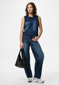 Jeans utan ärmar och vida jeans, båda i mörkblått med vita utsmyckningar. Modellen håller en svart handväska och bär silverfärgade sneakers.