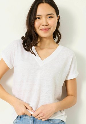 Młoda kobieta z ciemnymi włosami do ramion, ubrana w białą koszulkę z dekoltem w serek i niebieskie dżinsy, lekko uśmiechająca się na jednolitym tle.