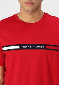 Camiseta de algodón roja con cuello redondo, con una banda gráfica en el pecho en tonos azul marino, blanco y rojo, y el nombre de la marca "TOMMY HILFIGER" en blanco.