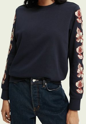 Sudadera azul marino con mangas largas que presenta un bordado floral rojo a lo largo de ambos brazos, combinada con jeans de tiro alto en azul oscuro.