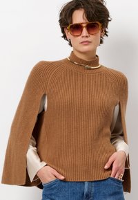Bruine geribbelde trui met hoge kraag en capesachtige mouwen; gelaagd over een lichtgekleurde shirt. Opvallende gouden hardwareaccent.