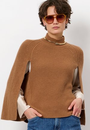 Bruine geribbelde trui met hoge kraag en capesachtige mouwen; gelaagd over een lichtgekleurde shirt. Opvallende gouden hardwareaccent.