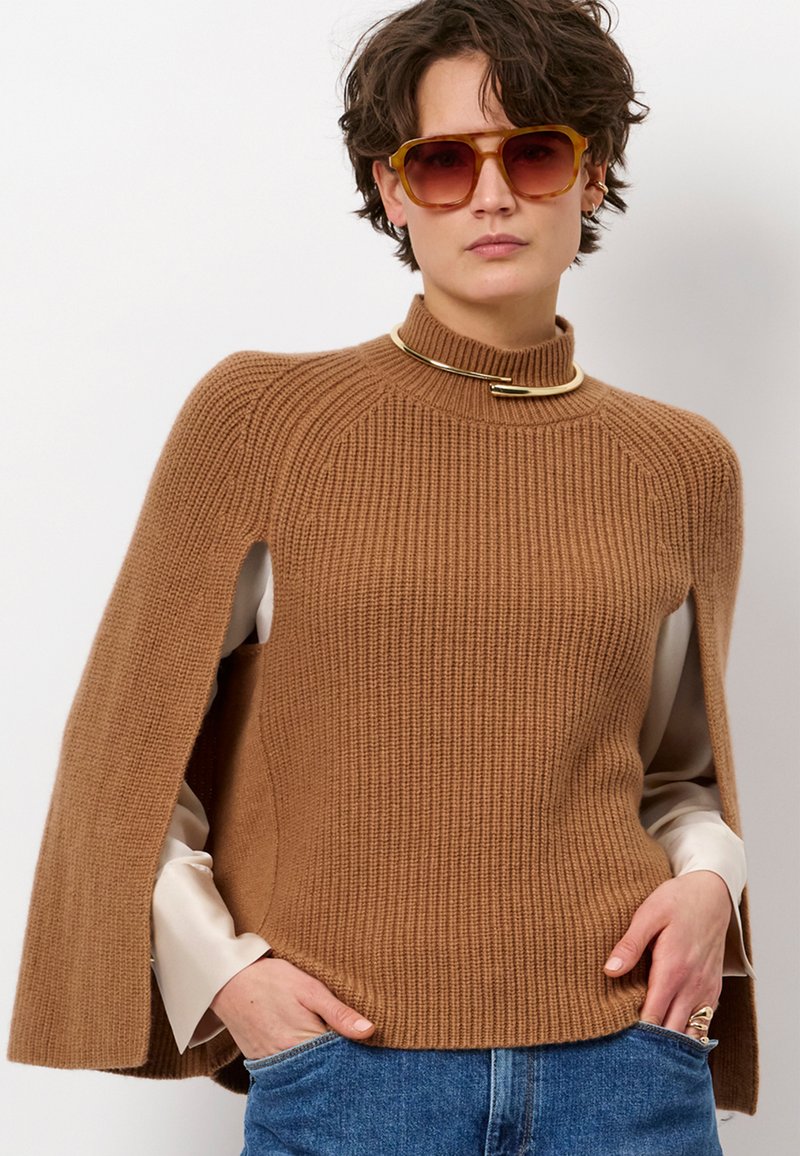 Bruine geribbelde trui met hoge kraag en capesachtige mouwen; gelaagd over een lichtgekleurde shirt. Opvallende gouden hardwareaccent.