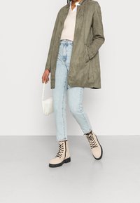 Persona che indossa jeans azzurri, stivali beige con lacci, cappotto verde oliva, tiene in mano una piccola borsa bianca con tracolla a catena, in piedi su un pavimento chiaro.