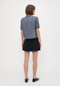 Korte ermer, cropped genser i flerfarget strikk med en subtil glans, kombinert med et svart skjørt, vist fra baksiden. Svarte slip-on sko.