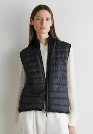 Frau trägt eine schwarze gesteppte ärmellose Weste über einer weißen langärmeligen Bluse und steht vor einer einfarbigen hellen Wand.