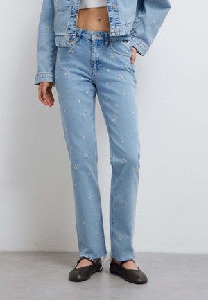 Jeans di denim azzurro chiaro con ricamo floreale bianco piccolo, indossati con giacca cropped di denim abbinata e ballerine nere.