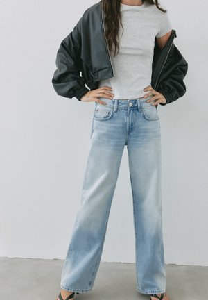 Femme portant un jean large bleu clair, un t-shirt blanc ajusté et une veste en cuir noire drapée sur les épaules, debout les mains sur les hanches.