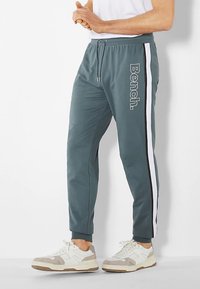 Sweatpants in dark teal fabric with a drawstring waist, side white stripe, and "Bench." logo printed along the left leg; cuffed ankles.
