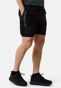 Zwarte sportbroek met een gestructureerde tailleband, voorzien van reflecterende "SWEDEMOUNT" tekst aan de zijkant. Gecombineerd met zwarte sneakers en sokken.