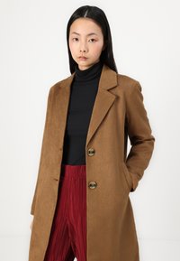 Brown suede coat with notched lapels, two buttons, and side pockets, worn over a black turtleneck and red pleated pants.