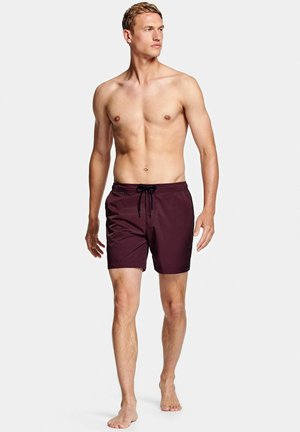 Man zonder shirt, blootsvoets lopend, draagt donkerrode zwembroek met zwarte trekkoord, tegen een effen lichte achtergrond.