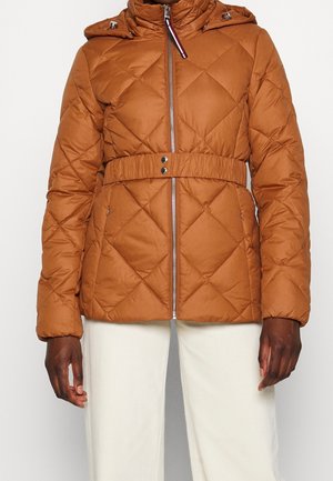 Veste matelassée marron avec capuche et ceinture à boutons-pression portée par une personne à la peau foncée et pantalon blanc sur fond uni.