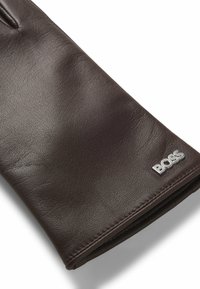Brun läderhandske med en slät struktur, prydd med en silverfärgad BOSS-logotyp. Klassisk design, elegant form, inga synliga mönster.
