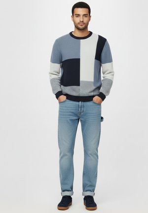 Man staat met de handen in de zakken, gekleed in een blauw, grijs en zwart kleurblok trui, lichtblauwe jeans en marineblauwe sneakers.