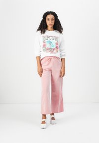Maglione bianco con grafica colorata "Ripcurl"; pantaloni a gamba larga a righe rosa; scarpe slip-on bianche. Outfit casual con vestibilità rilassata.