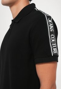 Black polo shirt featuring a textured fabric, two buttons on the collar, and white accent branding along the shoulder. Short sleeves.