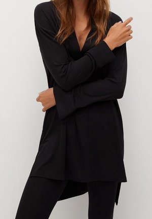Femme portant une tunique noire à manches longues avec un col en V, associée à des leggings noirs, posant les bras croisés devant un fond uni.