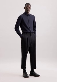 Camicia blu navy con bottoni e colletto, abbinata a pantaloni neri plissettati e scarpe di pelle nere. Tessuto liscio con una vestibilità su misura.