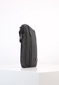 Borsa a tracolla nera, slim, in posizione verticale su una superficie bianca, con due scomparti zip visibili e materiale dalla texture liscia.