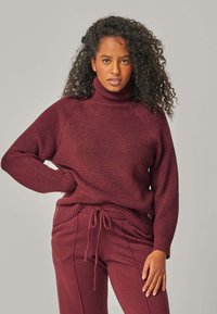 Frau mit lockigem Haar, die einen passenden burgunderroten Rollkragenpullover und eine Hose mit Kordelzug trägt, posiert mit einer Hand in der Hüfte vor einem schlichten Hintergrund.