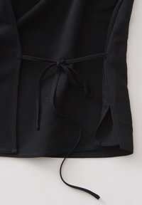 Haut enveloppant noir en tissu avec fermeture à nouer, fentes sur les côtés et un fini doux et texturé. Conçu pour un ajustement décontracté et un style sans effort.