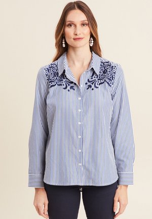 Gestreepte blouse met blauwe en witte verticale lijnen, voorzien van geborduurde bloemaccenten in marineblauw op de schouders. Knopsluiting aan de voorkant en manchetten met omslagen.