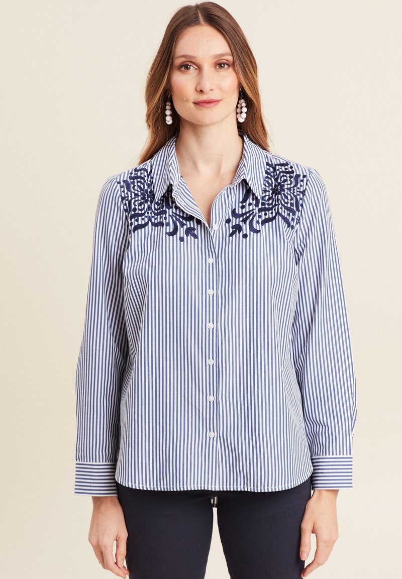 Blouse rayée avec des lignes verticales bleues et blanches, ornée d'accents floraux brodés en marine sur les épaules. Fermeture à boutons sur le devant et manches à revers.