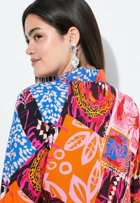 Kleurrijke blouse met een patchworkontwerp met levendige patronen, waaronder flora, dierenprints en abstracte vormen. Zilveren statement oorbellen.