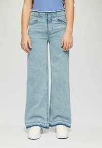 Jeans de mezclilla de color azul claro, de pierna ancha, con cintura alta, dos bolsillos frontales y detalles de dobladillo deshilachado, combinados con zapatillas blancas.