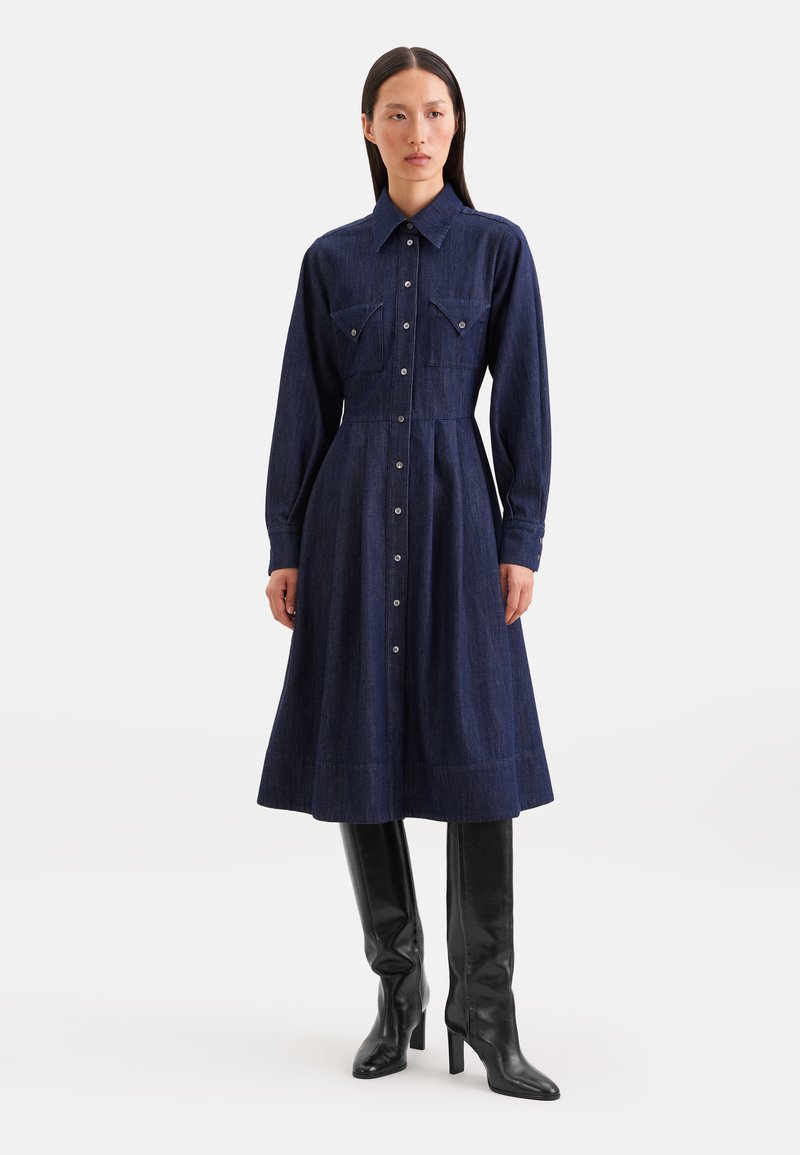 Femme portant une robe midi en denim foncé à manches longues boutonnée et des bottes noires à talons hautes jusqu'aux genoux, sur un fond blanc uni.
