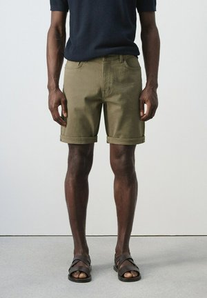 Hombre con pantalones cortos de color verde oliva con dobladillo enrollado, camisa negra de manga corta y sandalias negras, de pie sobre un suelo gris con fondo blanco.