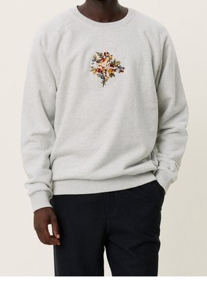 Homme portant un sweat-shirt gris clair avec une broderie florale multicolore sur la poitrine et un pantalon noir, debout avec une main dans la poche.