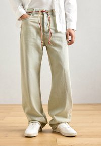 Light green wide-leg jeans with a drawstring waist, featuring contrasting colored ties. Worn with white sneakers on a wooden floor.