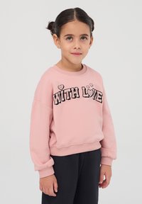 Felpa rosa con la scritta "WITH LOVE" in nero, adornata con cuori, polsini a costine e un tessuto morbido e testurizzato. Indossata con pantaloni scuri.