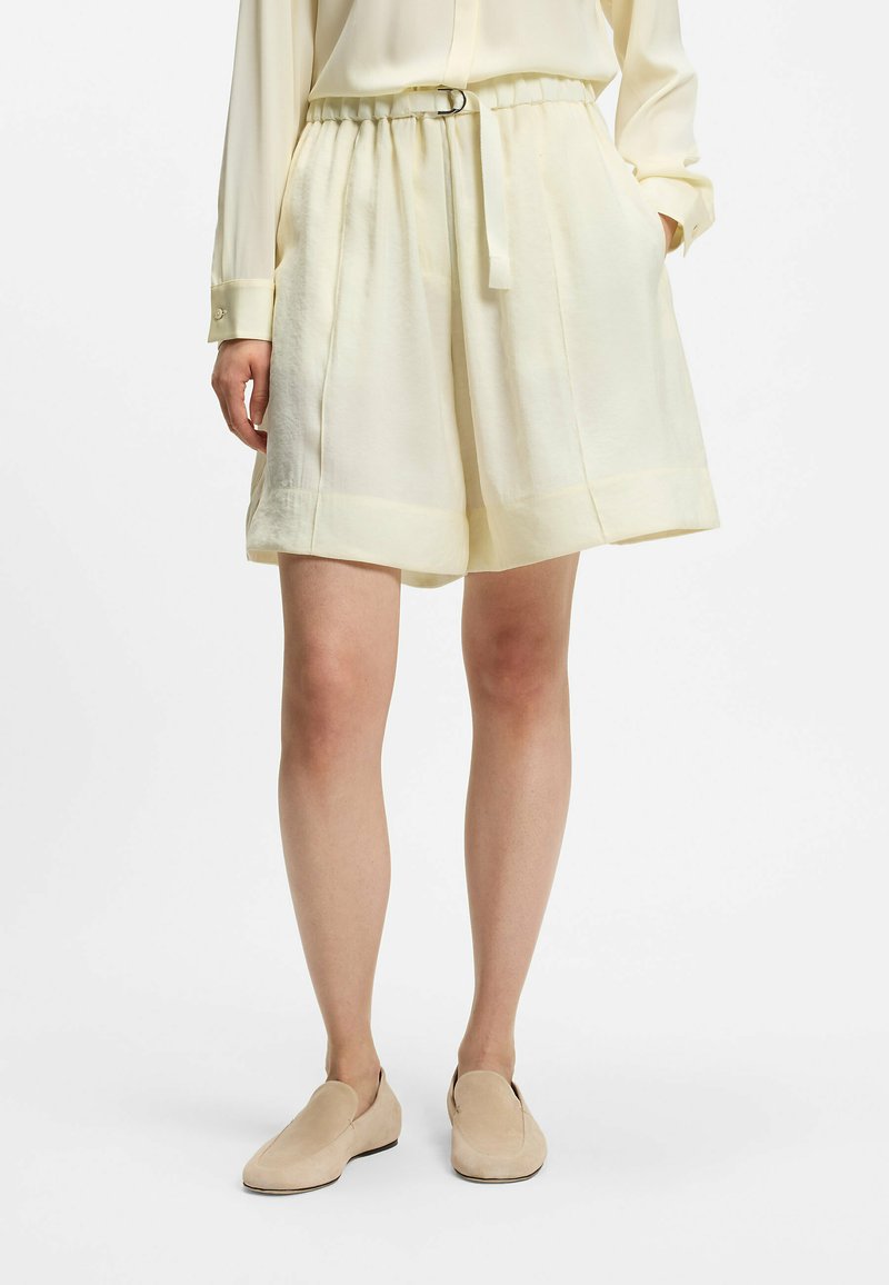 Femme portant un short ample couleur crème avec ceinture, un chemisier assorti et des chaussures beige slip-on, debout devant un fond blanc.