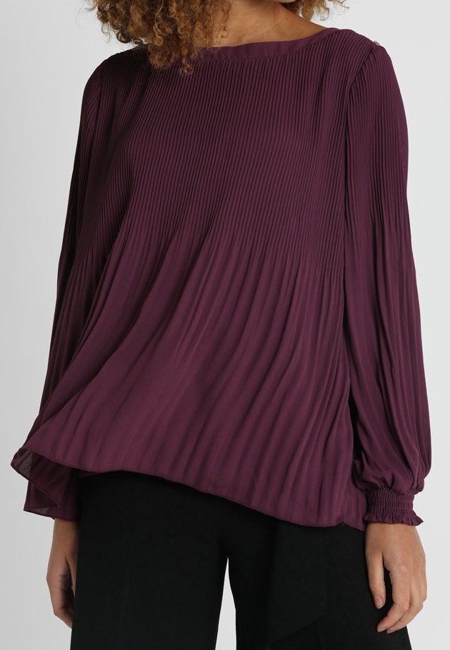 Kvinna i en långärmad, veckad, mörk lila blus tillsammans med svarta byxor, som visar midja och armar.