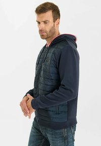 Sudadera híbrida azul marino con parte superior acolchada, puños y dobladillo acanalados. El cuello tiene acentos en rojo y blanco; bolsillos con cremallera en la parte frontal.