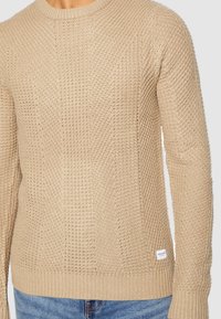 Beige neulepaita, jossa on teksturoitu kuosi, pyöreä kaula-aukko, resorit hihansuissa ja helmassa, sekä pieni etiketti vasemmassa alareunassa.