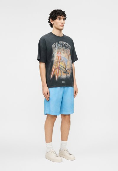 Jeune homme portant un t-shirt graphique noir délavé, un short bleu clair, des chaussettes blanches et des baskets beiges, debout devant un fond uni.
